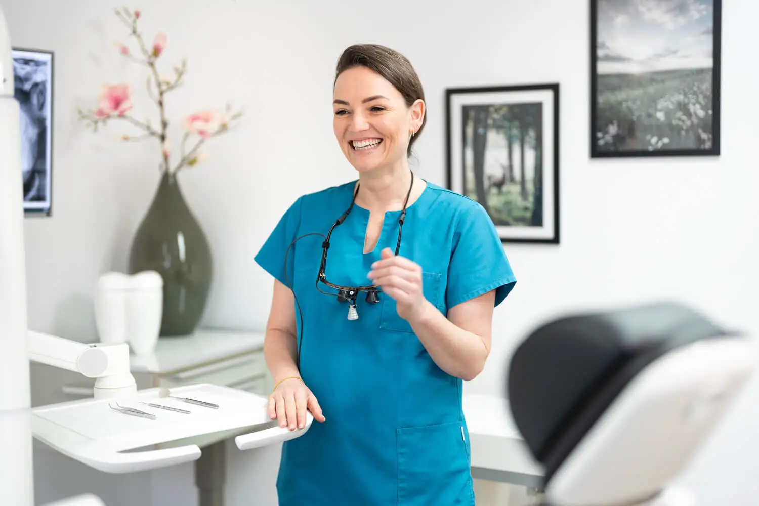 Eine Frau mit braunen Haaren trägt einen blauen Arztkittel. Sie lächelt und steht in einer Klinikumgebung mit hellen Wänden und medizinischer Ausrüstung im Hintergrund.