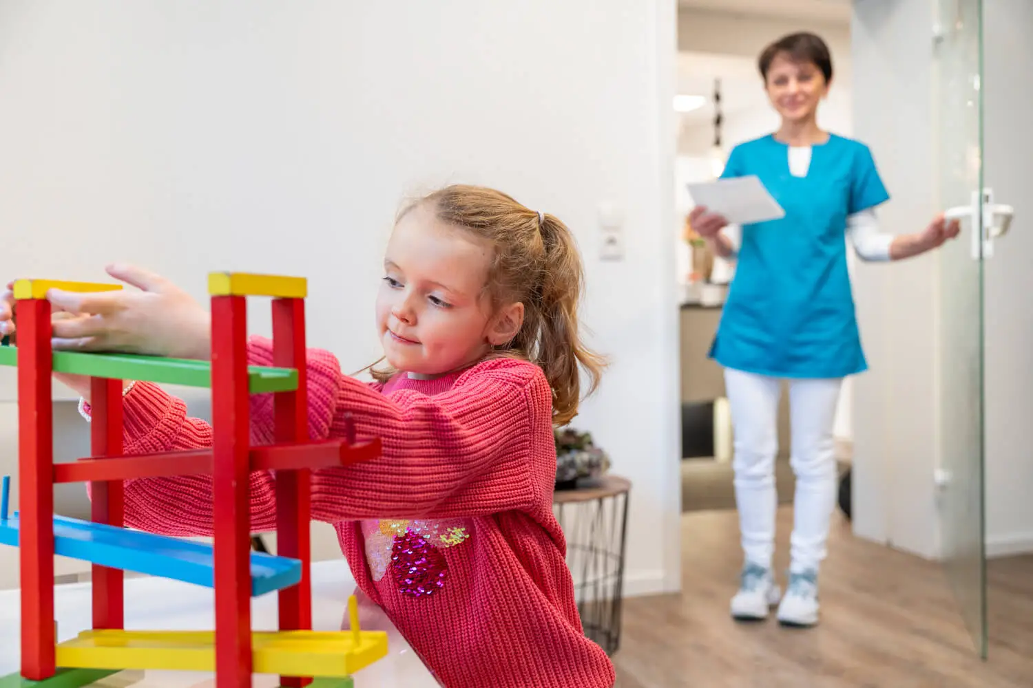 Ein junges Mädchen in einem rosa Pullover mit verspieltem Muster baut eine bunte Spielzeugkonstruktion aus Holz zusammen. Im Hintergrund lächelt eine Frau in einem blauen OP-Outfit, die ein Klemmbrett hält und in einem hellen, modernen Raum auf das Mädchen zugeht.