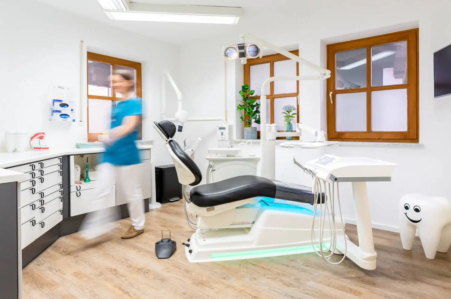 Moderne Zahnklinik mit einem Zahnarztstuhl in der Mitte, einer verschwommenen Figur eines Arztes in blauem Kittel, Schränken mit Zahnarztinstrumenten und einer zahnförmigen Dekoration in der Ecke. Der Raum hat weiße Wände, Holzböden und Fenster mit Holzrahmen.
