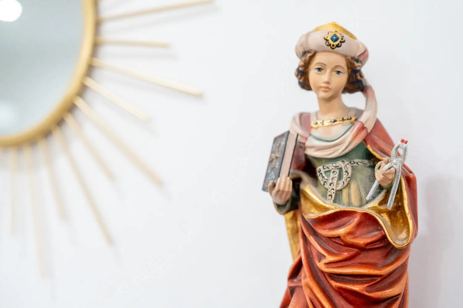 Vor einem weißen Hintergrund steht eine detailreiche Statue eines Heiligen in mittelalterlicher Kleidung mit einer Krone, der in einer Hand ein Buch und in der anderen eine Schreibfeder hält. Im Hintergrund ist teilweise ein dekorativer Sonnenspiegel zu sehen.