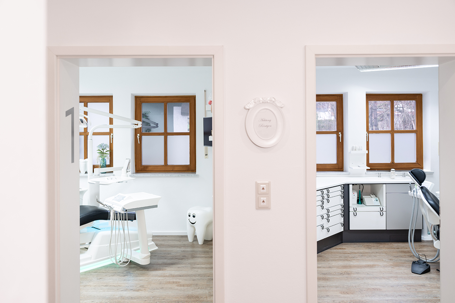 Eine moderne Zahnklinik mit zwei Behandlungsräumen, die durch Türen sichtbar sind. Der linke Raum verfügt über einen Zahnarztstuhl und Geräte, während der rechte Raum über Schränke und eine Theke verfügt. Beide Räume sind sauber, gut beleuchtet und haben Fenster mit Milchglas. Die Wände sind weiß und die Böden aus Holz.