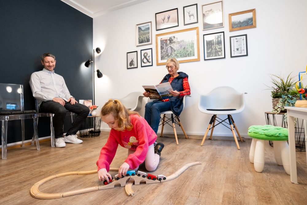 Ein Kind spielt mit einer Holzeisenbahn auf dem Boden eines Wartezimmers. Eine ältere Frau sitzt auf einem Stuhl und liest eine Zeitschrift, während ein Mann daneben sitzt und lächelt. Der Raum ist mit gerahmten Fotos und minimalistischen Möbeln dekoriert.