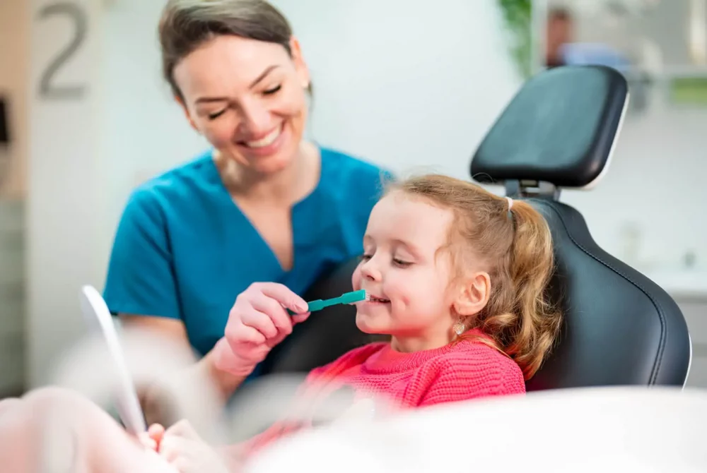 Eine lächelnde Frau in einem blauen OP-Kittel hilft einem jungen Mädchen in einem rosa Pullover beim Zähneputzen, während sie im Zahnarztstuhl sitzt. Das Kind hält einen kleinen Spiegel und sorgt so für eine freundliche und ermutigende Zahnpflegeszene.